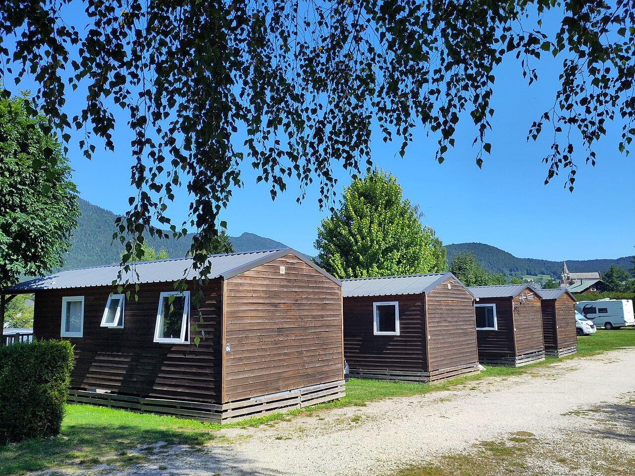 Quatre petits hébergements locatifs à bardage de bois sombre et toit métallique, alignés le long d'une allée de camping